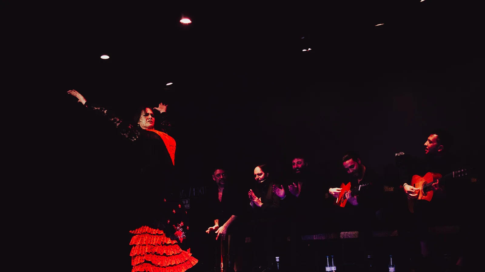 A flamenco cuadro on stage: a dancer in a red ruffled dress mid-performance, with guitarists and singers clapping palmas beside her